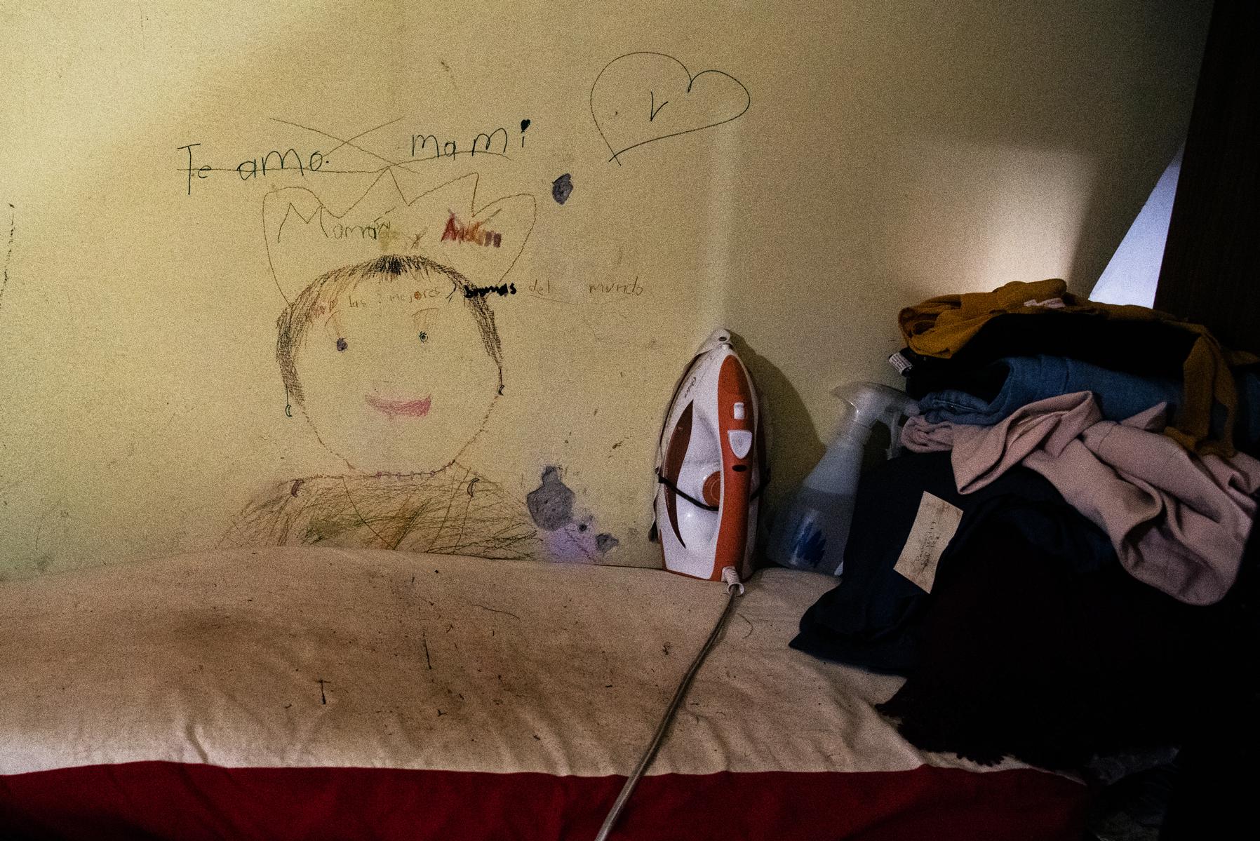 Burro para planchar con una plancha, ropa doblada y un dibujo que dice "Te amo mami" ilustrado sobre la pared