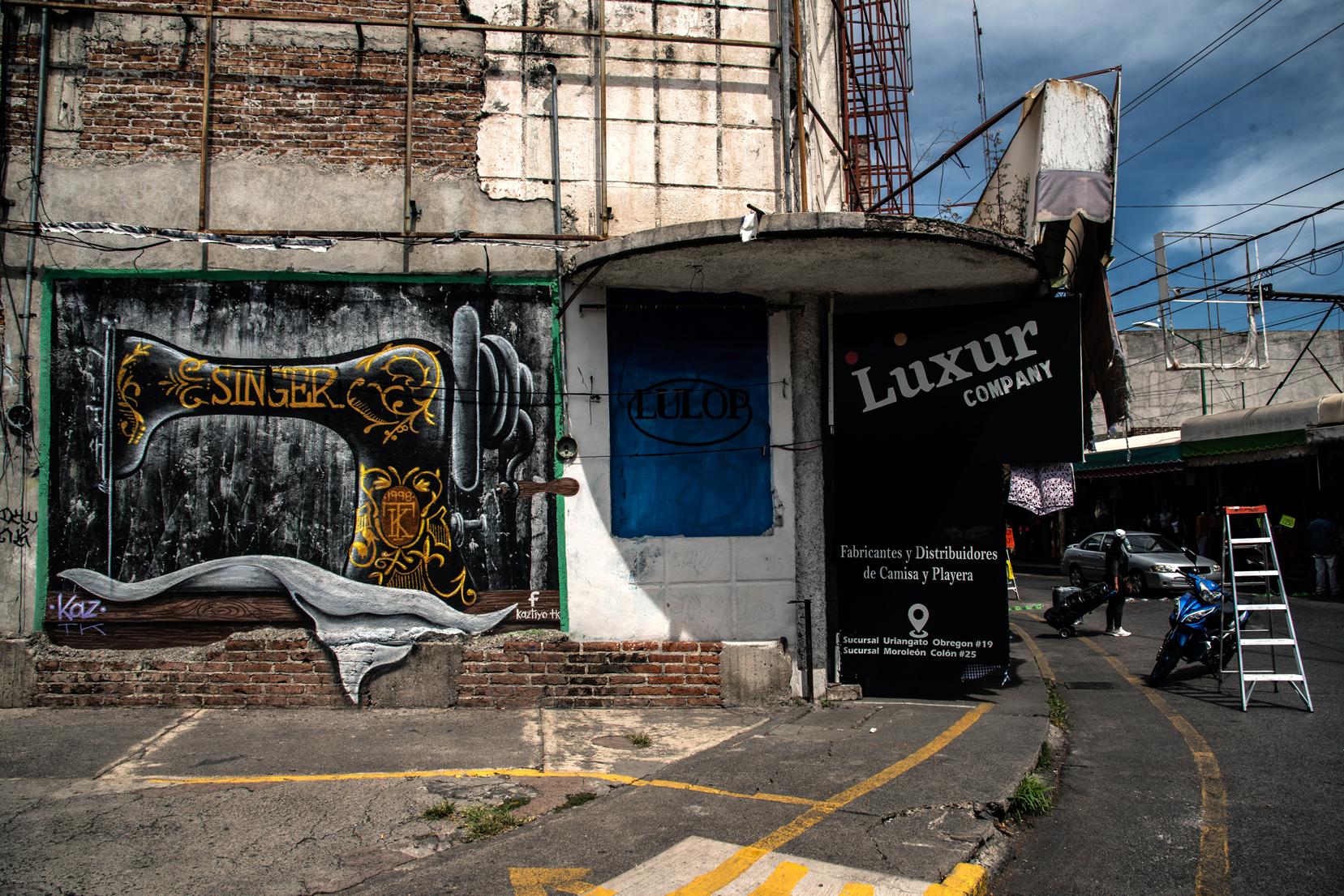 Fachada de un edificio con un mural de una máquina 'Singer' y un local de fabricación y distribución de camisas y playeras
